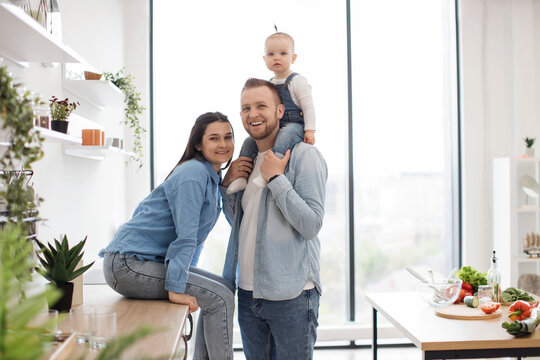Portrait of cheerful mom sitting on countertop while snuggling to strong dad with kid on shoulders in dining room. Joyful family of three enjoying pleasant pastime spent in spacious kitchen interior.