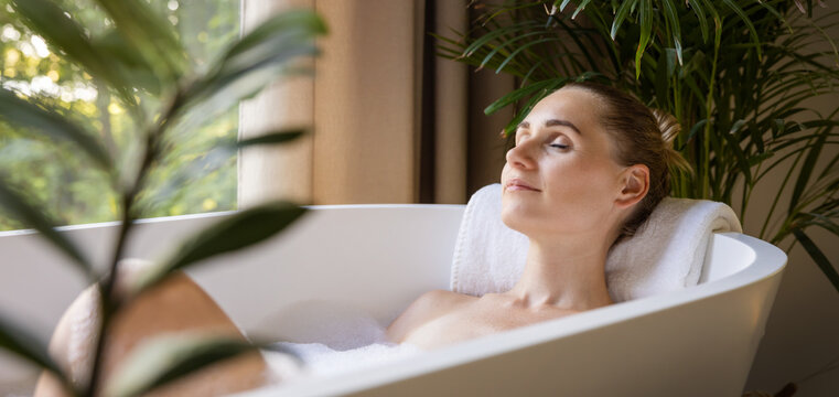 woman relaxing in bathtub with eyes closed in bathroom with green tropical plants. spa treatment, body and skin care. banner with copy space - Powered by Adobe