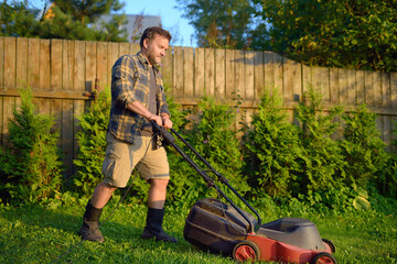 Mature man gardener cutting grass in his backyard with lawn mower on summer sunny day. Lawnmower...