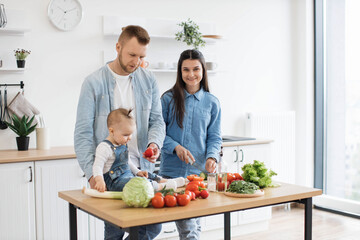 Handsome father in denim shirt giving tomato to little daughter while pretty mother mastering knife skills at kitchen table. Little infant learning colors and forms while relishing family activities.
