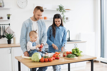 Handsome father in denim shirt giving tomato to little daughter while pretty mother mastering knife skills at kitchen table. Little infant learning colors and forms while relishing family activities.