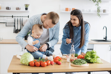 Funny little girl in cozy outfit sitting on dining table while caring young mother holding bite-sized slice of tomato. Mindful parents motivating infant daughter to making healthier food choices.