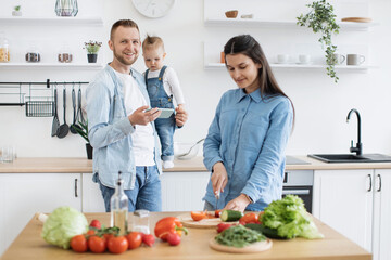 Smiling blond man using cell phone while standing near countertop with infant girl in home kitchen. Loving mother utilizing clean raw ingredients for homemade dishes to please entire family.