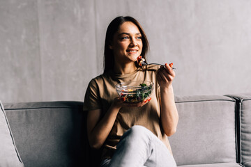 Happy young woman relaxing on the sofa eating salad in her living room