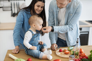 Fototapeta na wymiar Close up view of beautiful little girl in cozy outfit eating tasty slice held by caring father while mom smiling behind. Young couple weaning baby girl by giving solid food while making vegan meals.