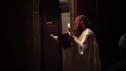 Priest stands with a lighted candle and the Bible during Easter ceremony in the Ortodox church, Easter tradition of knocking with wooden hamer on the church door