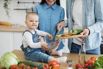 Close up view of funny baby girl making angry face with bit lower lip while holding salad bowl with mother's assistance. Helpful daughter being involved in process of salad preparation in kitchen.