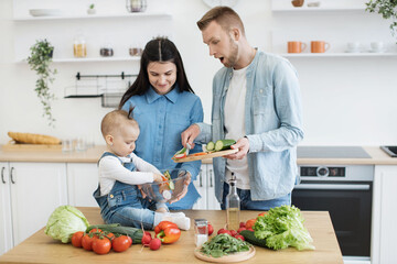 Shocked father holding cutting board with sliced cucumber while sweet baby girl mixing salad with hands in glass bowl. Curious kid exploring texture and shapes of colorful punch of salad ingredients.