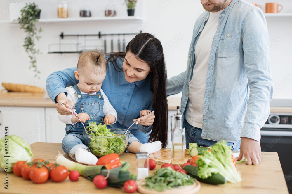 Sticker Cropped view of male in denim clothes leaning on wooden table covered with products and kitchenware. Attractive mother using spoons for salad mixture while active kid checking greens in bowl.