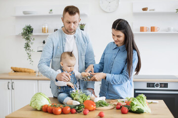 Beautiful brunette lady pouring liquid into bowl with chopped vegetables and greens while baby girl helping with bottle. Friendly young family sharing and bonding while working together in kitchen.