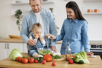 Affectionate young family with little infant having fun together while cooking natural balanced food in kitchen. Happy smiling parents enjoying weekend time with small child. Concept of healthy diet.