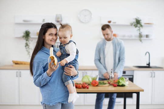 Cheerful caucasian mother proposing baby tasty banana while father making nutritious dinner for whole family on blur background. Little girl getting acquainted with complementary foods.