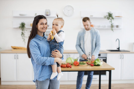Charming lady with thick long hair holding baby on hand and smiling at camera while infant enjoying delicious banana indoors. Mother and daughter waiting for dinner while daddy making vegetable salad.