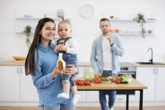 Precious little cutie with ponytail eating organic banana while sitting on mommy's hands. Responsible father cutting and cooking vegetables for lunch on wooden table in white kitchen.