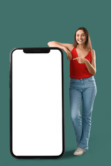 Happy young woman pointing at big mobile phone with blank screen on green background. Banner for design