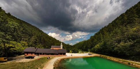 Scenic shot of a building and a pond in the middle of forest and mountain valley under a cloudy sky