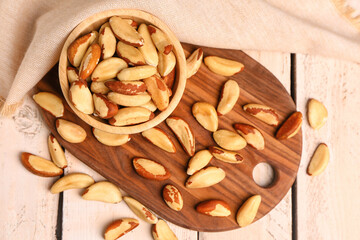 Cutting board with bowl of tasty Brazil nuts on light wooden background