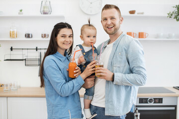 Portrait of beaming parents and adorable baby girl standing with nutritious juice glasses and looking at camera. Pretty little girl sipping beverage while sitting on father's hand at home.