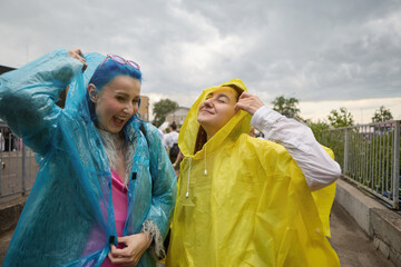 Two young lesbian females in raincoats having fun. Portrait of a couple of diverse LGBT persons laughing outdoor in a rainy day