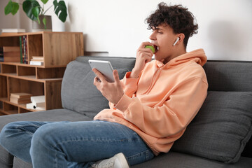 Male student with tablet computer eating apple on sofa at home