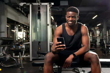 young athletic african american man sits in dark gym and uses smartphone, sports guy types online on the phone
