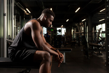 young athletic african american man sits in dark gym and thinks, pensive athletic guy rests and looks forward