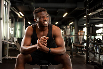 Obraz na płótnie Canvas young athletic african american man smiling in dark gym and daydreaming, athletic guy resting and looking away