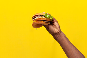 african american male hand holds and shows tasty burger on yellow isolated background, close-up of...