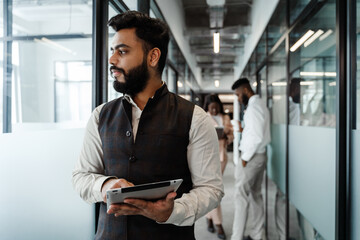 Young indian man in suit holding tablet computer at office corridor