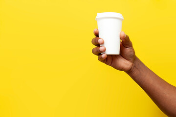 african american man's hand holds and offers white paper cup of coffee on yellow isolated background