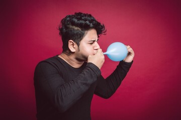 Young Indian man blowing up a blue balloon, preparing for a party over a red background