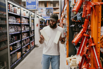 Young handsome man with a beard in casual clothes in a construction hypermarket chooses tools - obrazy, fototapety, plakaty