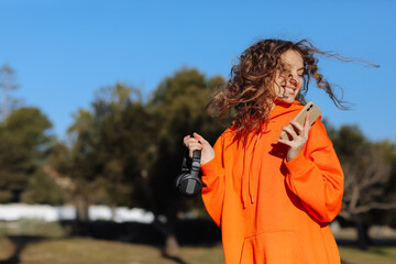 Smiling young woman with curly hair wearing bright orange sweatshirt holds headphones for music,...
