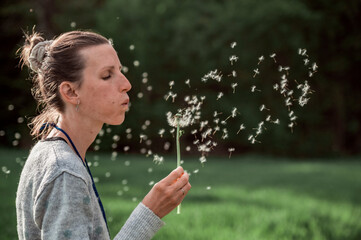 Young woman blowing dandelion bulb with seeds floating around her