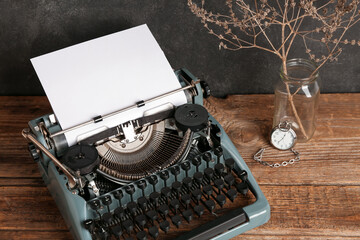 Vintage typewriter, pocket watch and vase with dried flowers on wooden table near black wall