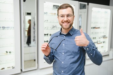 In Optics Shop. Portrait of male client holding and wearing different spectacles, choosing and trying on new glasses at optical store. Man picking frame for vision correction, closeup.