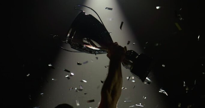 Silhouette of unrecognizable Caucasian male raising a trophy above head against bright light and falling confetti. Super slow motion, shot on RED cinema camera