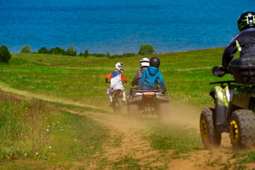 A group of tourists riding motorcycles and quad bikes on a field road.  In the background, the shore of a body of water.