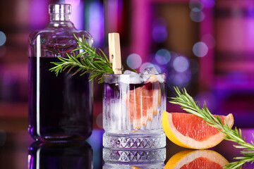 Glass of purple gin and tonic with bottle, grapefruit and rosemary on table in bar against blurred...