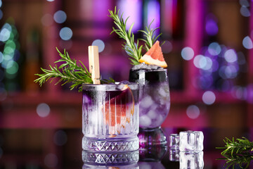 Glasses of purple gin and tonic with ice cubes, grapefruit and rosemary on table in bar against blurred background