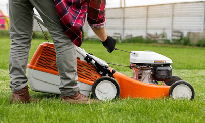 Unrecognisable gardener man in protective gloves starts the lawnmower before cutting green grass lawn in his backyard. Man with motorised lawnmower cares for landscaping lawn