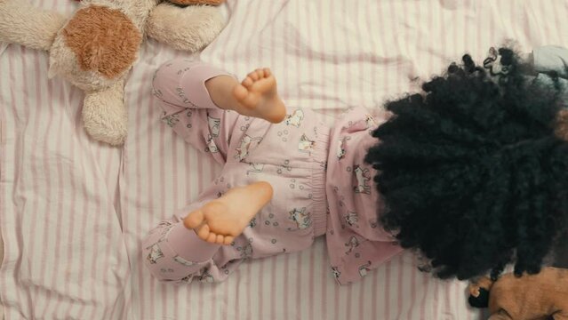 Top view of a cute child girl with curly hair lying and reading a book on bed in the room