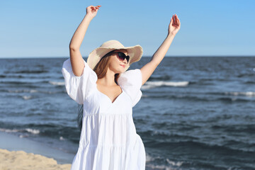 Fototapeta na wymiar Happy smiling woman in free happiness bliss on ocean beach standing with a hat, sunglasses, and rasing hands. Portrait of a multicultural female model in white summer dress enjoying nature during trav