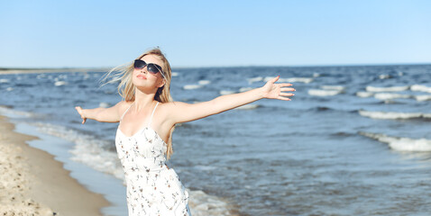 Fototapeta na wymiar Happy blonde beautiful woman on the ocean beach standing in a white summer dress and sun glasses, open arms.