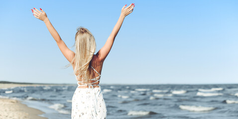 Happy blonde beautiful woman on the ocean beach standing in a white summer dress and glasses, raising hands.