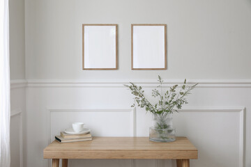 Fototapeta na wymiar Two empty vertical picture frame mockups hanging on wall. Cup of coffee, books. Wooden desk, table. Vase, green grasses and cow parsley. Minimal working space, home office. Elegant Scandi interior.