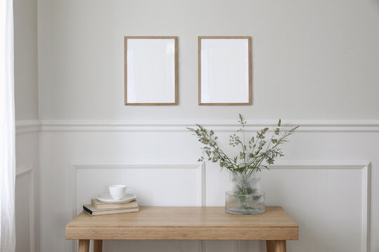 Two empty vertical picture frame mockups hanging on wall. Cup of coffee, books. Wooden desk, table. Vase, green grasses and cow parsley. Minimal working space, home office. Elegant Scandi interior.