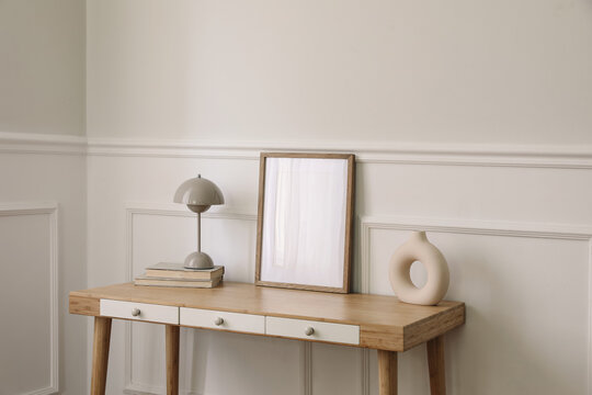 Modern living room, home office. Trendy round vase, beige lamp and old books. Blank picture frame mockup. Bamboo table with white drawers. Minimal Scandi boho interior, home. Beige wall background.