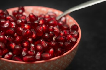 Closeup of a bowl full of red pomegranate juicy seeds, served with a spoon, on the black background