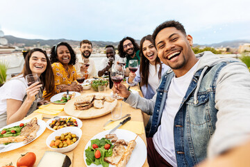 Happy group of people toasting red wine at eatery barbecue lunch party. Multiracial friends taking selfie portrait together enjoying food and drink at summer celebration. Friendship concept